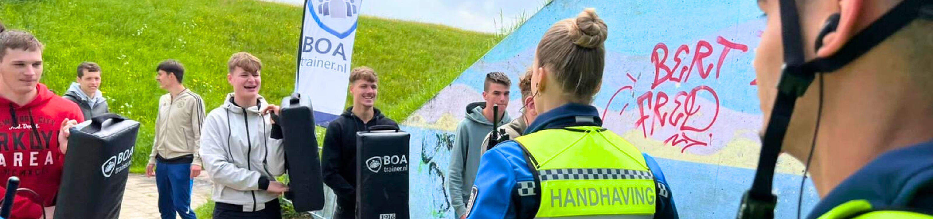 A group of young people and a uniformed officer engage in outdoor training with padded equipment near a graffiti-covered wall.