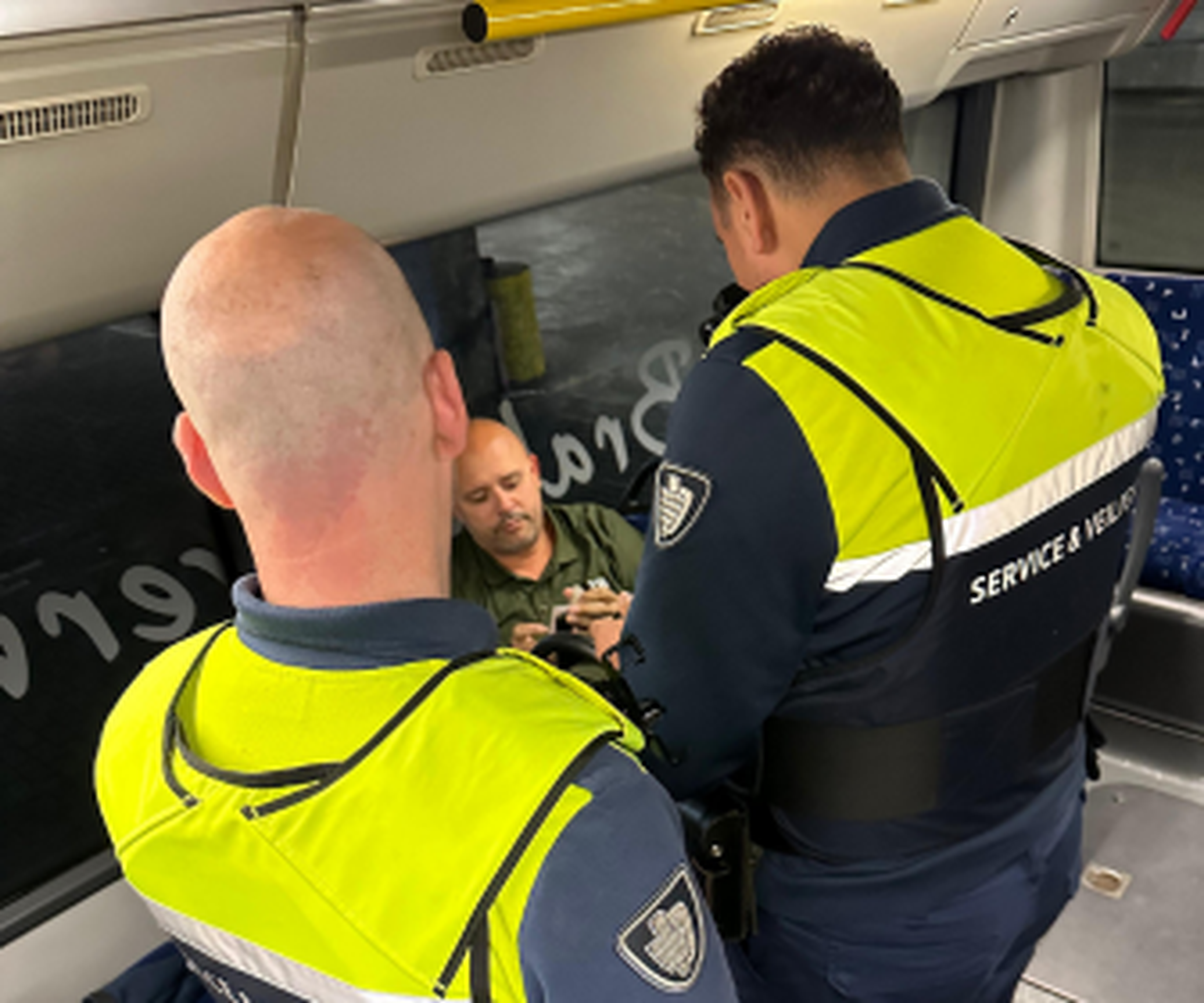 Two uniformed security personnel check a seated man's documents on a bus.