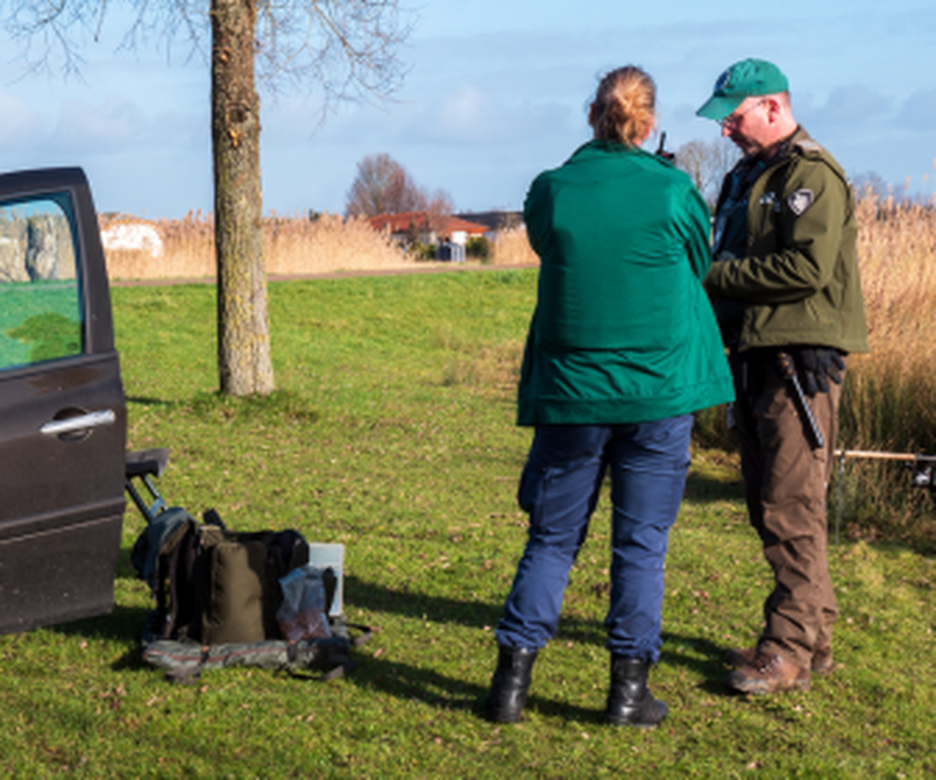 Two people in outdoor gear stand talking near a parked vehicle on grassy terrain, with trees and reeds in the background.